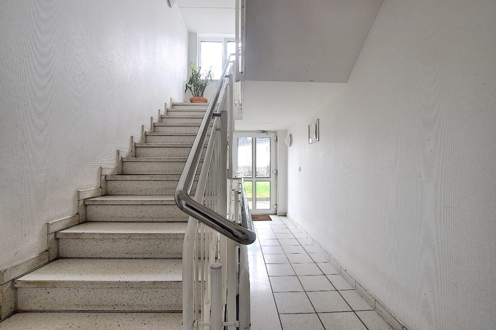 Cage d'escalier d'un immeuble collectif aux murs clairs et au sol carrelé, vue sur la porte d'entrée par-delà la rampe en acier inoxydable