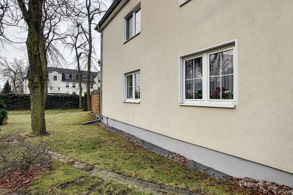 Vue latérale d'une maison individuelle au crépi beige, dotée de deux fenêtres à croisillons blanches et d'une pelouse dans le jardin de devant