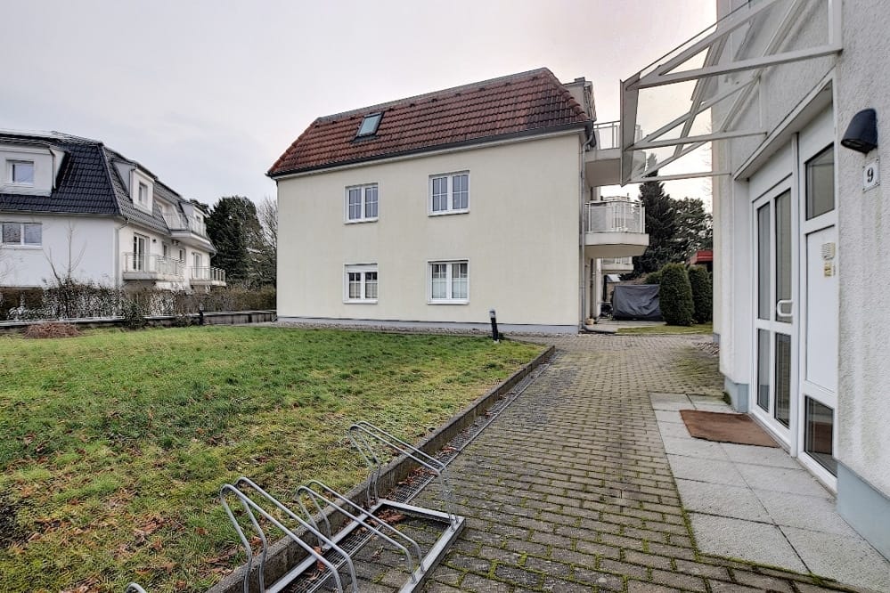 Vue de face d'un immeuble collectif de deux étages, avec un toit en pente rouge, un balcon et un jardin devant la maison équipé d'un porte-vélos