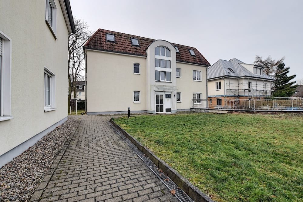 Immeuble collectif de trois étages avec un crépi clair, un toit en tuiles rouges, une fenêtre en arc au centre et un jardin devant la maison avec une pelouse