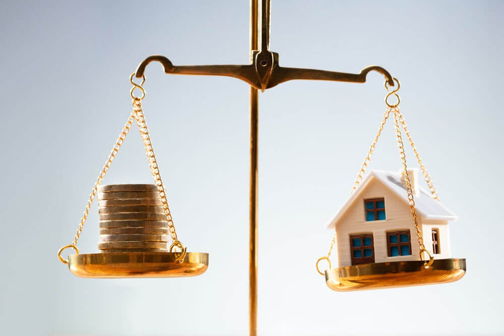 Frontal close-up of a golden scale with a stack of coins on the left pan and a white model house on the right pan