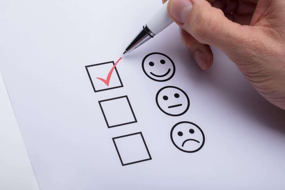Nahaufnahme Hand mit Stift setzt roten Haken in oberstes Kästchen eines Smiley-Bewertungsbogens von oben