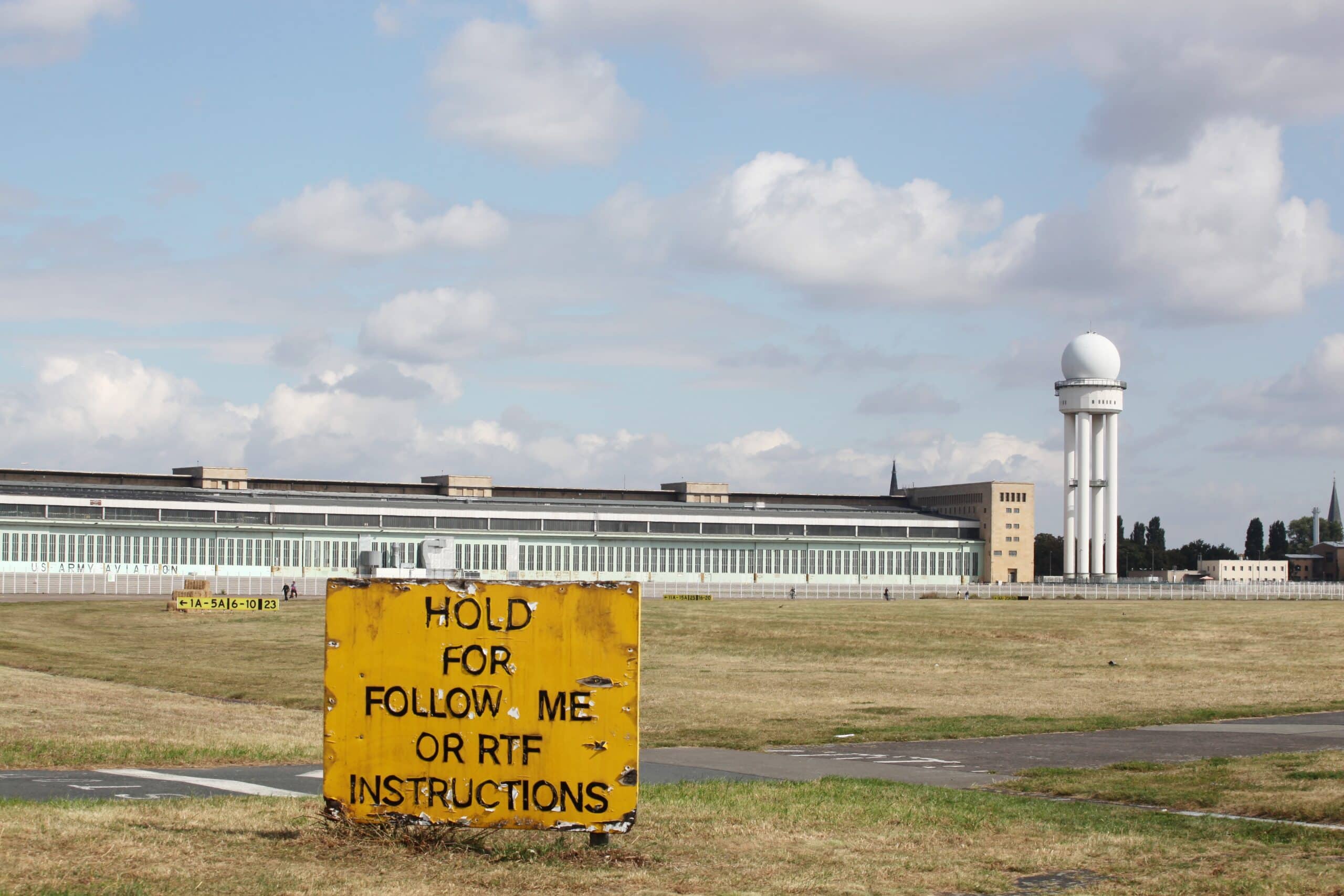Vue de face de l'ancien hangar à avions avec l'inscription US Army Aviation et la tour radar ronde derrière la pelouse libre