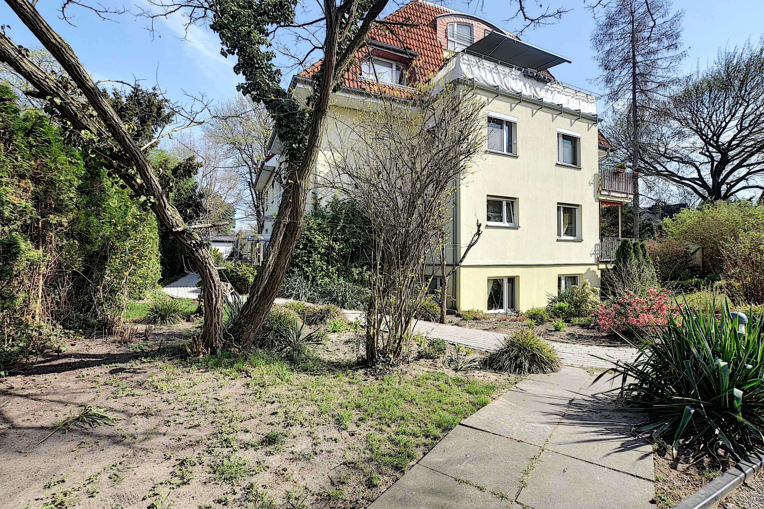 Mehrfamilienhaus von vorne mit gelber Putzfassade, roter Dachgaube, seitlichem Balkon und gepflegtem Vorgarten