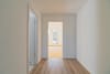 Innenansicht eines Wohnungsflurs mit Holzboden und Blick ins helle Wohnzimmer mit bodentiefen Fenstern und kleinem Balkon