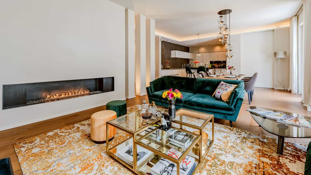 Offene Wohnküche in moderner Loftwohnung mit Wandkamin, grünem Samtsofa, goldfarbenem Couchtisch und Esstisch im Hintergrund