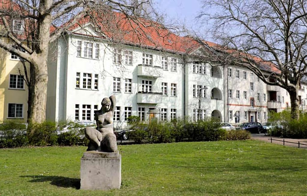 Dreigeschossiges Wohnhaus mit rotem Satteldach und Balkonfront aus leichter Perspektive davor Parkwiese mit Skulptur
