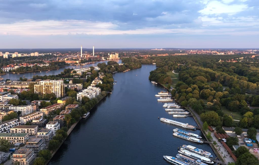 Luftaufnahme eines modernen Wohnkomplexes am Flussufer mit Balkonen und Bootsanleger neben Parklandschaft