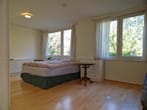 Vista interior do quarto a partir da perspetiva do corredor com cama de casal, janela grande, cortinas, cómoda e chão em parquet.