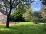 Jardim com relvado e terraço em frente à extensão envidraçada de uma casa unifamiliar à sombra de grandes árvores