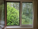 Vista de janela aberta para jardim verde com arbustos densos e relvado