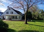 Vista do jardim de uma casa unifamiliar branca com um jardim de inverno envidraçado, uma fachada lateral com empena e uma grande área de relvado.