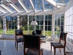 Vista interior de um jardim de inverno com teto de vidro, paredes de vidro, mesa redonda de vidro, cadeiras de madeira e vista para o jardim.