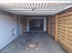 Garagem individual aberta, vista frontal com pavimento em calçada, parede de tijolo branca à esquerda, revestimento de ripas de madeira e prateleiras à direita