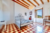 Schlafzimmer mit schmiedeeisernem Bett, sichtbaren Holzbalken, rot-weißem Kachelboden und offenem Fenster mit Bergblick
