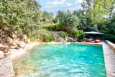 Frontaler Blick auf natürlichen Schwimmteich mit steiniger Einfassung Sonnenschirm und roten Sitzkissen im Wald