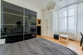 Blick ins Schlafzimmer mit Bett, spiegelndem Schiebetürenschrank, Holzdielen und Fenster mit Spitzenvorhängen