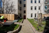 Innenhof eines Mehrfamilienhauses mit gepflastertem Weg, begrüntem Garten, Sitzbank und Hauseingang