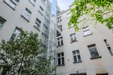 Innenhofansicht mehrgeschossiger Wohnhausfassade mit grauer Putzfassade und freistehendem Glasaufzugsturm