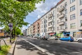 Straßenansicht von mehrgeschossigen Wohnhäusern mit Balkonen und Erdgeschossladen unter begrünten Bäumen