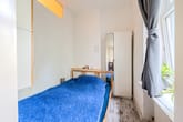 Innenraum eines kleinen Schlafzimmers mit Holzbett, blauem Überwurf, weißem Spiegelschrank und Vorhangfenster