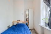 Schlafzimmer mit Einzelbett (Holzrahmen), blauer Decke, weißem Spiegelschrank und Fenster mit Topfpflanze