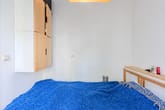 Innenansicht Einzelzimmer mit blauem Bettüberwurf wandhängendem Holz-Weiß-Schrank und Holzregal als Kopfteil