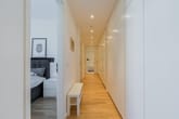 Blick in modernen Apartmentflur mit hellen Einbauschränken, Parkettboden, Deckenspots und Sitzbank neben Schlafzimmer