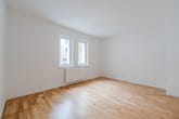 Vista frontal de uma sala de estar vazia com chão em parquet claro, duas janelas do chão ao teto e um radiador branco.