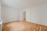 Vista interior de um apartamento com soalho de madeira claro, paredes brancas e porta aberta, fotografada do canto da sala