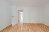 Quarto vazio com piso de madeira clara e porta aberta para o corredor, com uma fachada de vidro visível ao fundo.