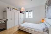 Vista interior de um quarto com cama de casal, um grande roupeiro branco, chão em parquet e janela com vista para a varanda.