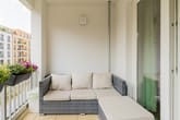 Varanda moderna de um apartamento com sofá de vime cinzento, almofadas, floreiras na varanda, fachada em estuque claro