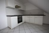 Apartamento em sótão com cozinha equipada em L com móveis superiores brancos, bancada preta, parede de azulejos e teto inclinado