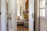 Durch offene weiße Türen Blick in helles Schlafzimmer mit Doppelbett, gemusterten Kissen, Holzbeistelltisch und Fenster