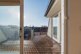 Balkon einer Dachgeschosswohnung innen durch offene Schiebetür mit Holzterrasse und Metallgeländer mit Blick auf Stadtdächer