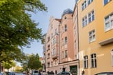 Straßenseitige Ansicht eines Altbau-Mehrfamilienhauses mit rundem Eckturm und Balkonen, begrünt durch Bäume im Vordergrund