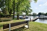 Park bench overlooking wooden pier and motorboats at green lake shore under deciduous trees