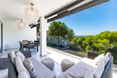 Moderner Appartementbalkon mit Lounge-Sofa, Esstisch und Glasgeländer vor Pinienhain und Meerblick