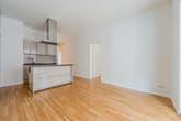 Offener Wohnbereich einer Neubauwohnung mit Kochinsel, Edelstahl-Dunstabzug und Parkettboden aus Eingangsansicht