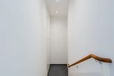 Treppenabgang im Wohnhaus mit schmalem weißen Flur, Holzhandlauf rechts und dunkelgrauen Bodenfliesen, Blick von oben