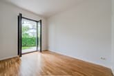 Blick aus leerem Wohnzimmer einer Erdgeschosswohnung mit Parkettboden durch bodentiefe Glastür in grünen Garten