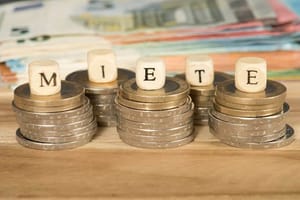 Close-up van gestapelde euromunten met houten letterblokjes waarop MIETE staat, vaag bankbiljet op de achtergrond