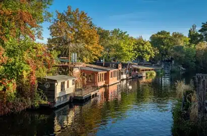 Blick auf mehrere Hausboote am baumgesäumten Flussufer mit Holzverkleidung und Sonnendeck