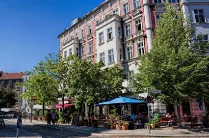 Eck-Gründerzeithaus in Seitenansicht mit Balkonen und Straßencafé unter Bäumen