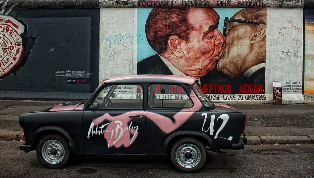 Frontalansicht bemalter Betonwand an der East Side Gallery mit Bruderkuss-Graffiti und parkendem schwarzen Trabant