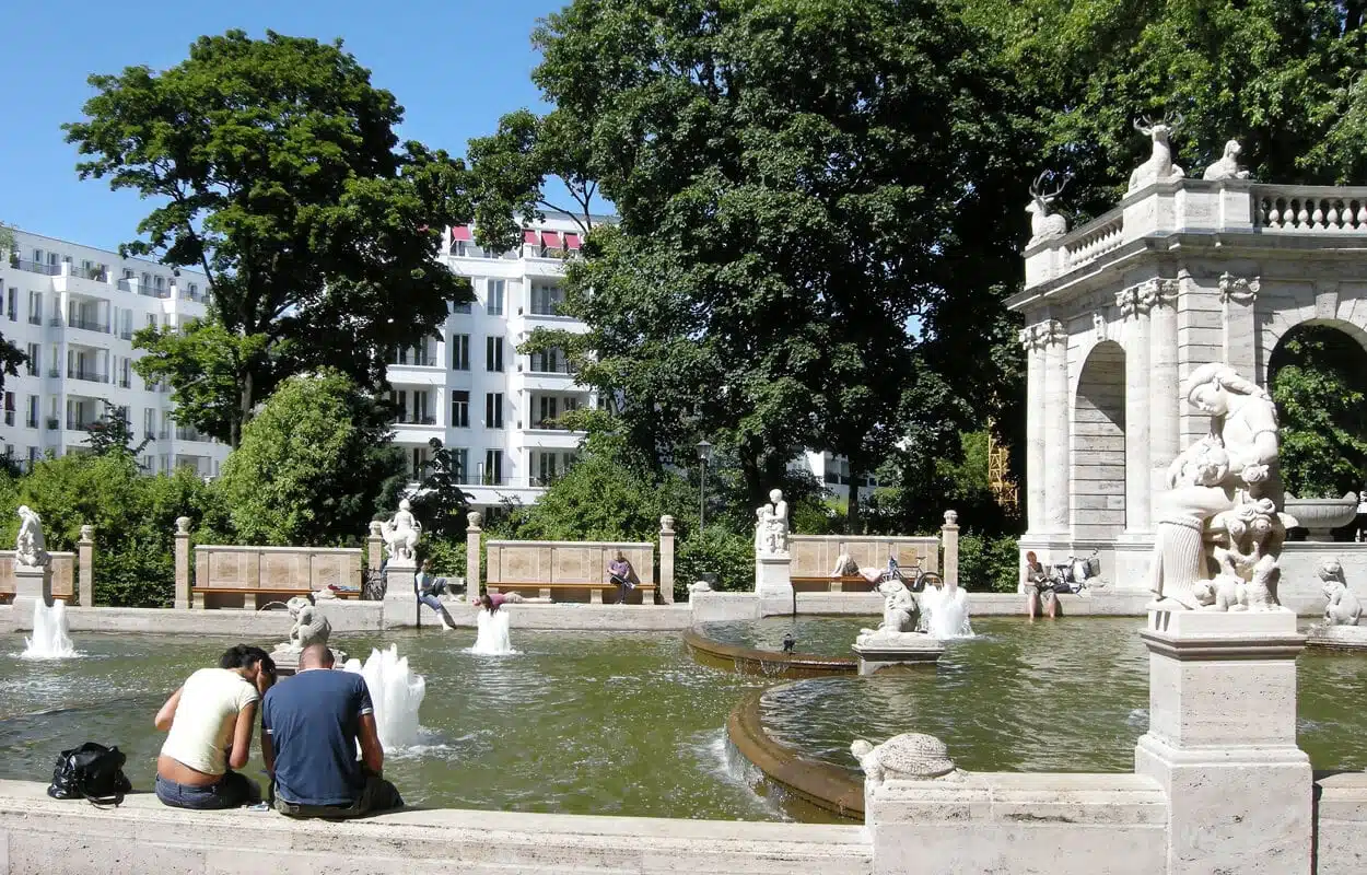 Frontalansicht eines weißen Mehrfamilienhauses mit Balkonen vor Springbrunnen und Parkvegetation