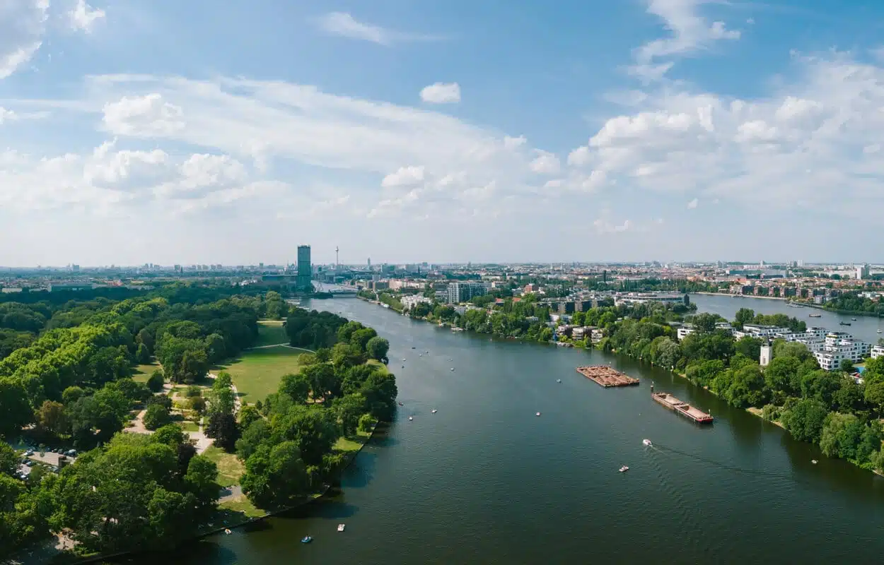 Vogelperspektive auf Flussufer mit Park links und moderner Wohnbebauung rechts mit Uferpromenade und Skylineblick