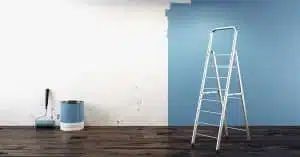 Aluminium-Leiter vor halb blau gestrichener Wand mit Farbroller und Farbeimer auf dunklem Holzdielenboden