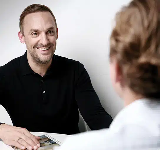 Makler sitzt gegenüber Kunde an Tisch mit Immobilienprospekt in Beratungsgespräch